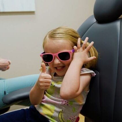 A young girl with pink glasses on smiling and giving a thumbs up in a pediatric office in Austin